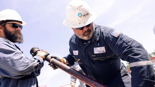 Two workers in Nine Energy hard hats and blue coveralls handling drilling pipe at an outdoor oilfield worksite